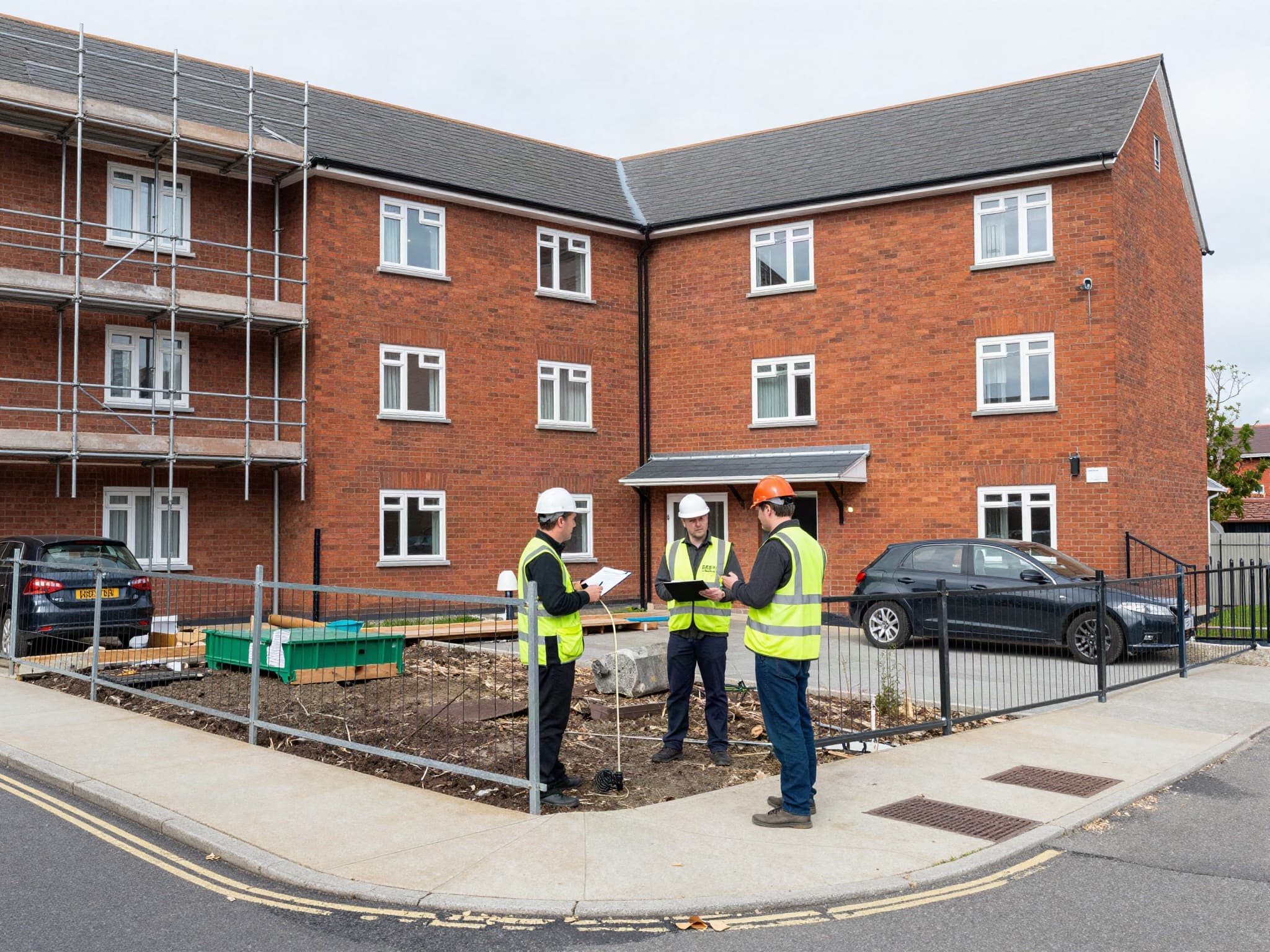 Three site managers in hard hats and hi-vis reviewing plans outside a brick residential block with scaffolding