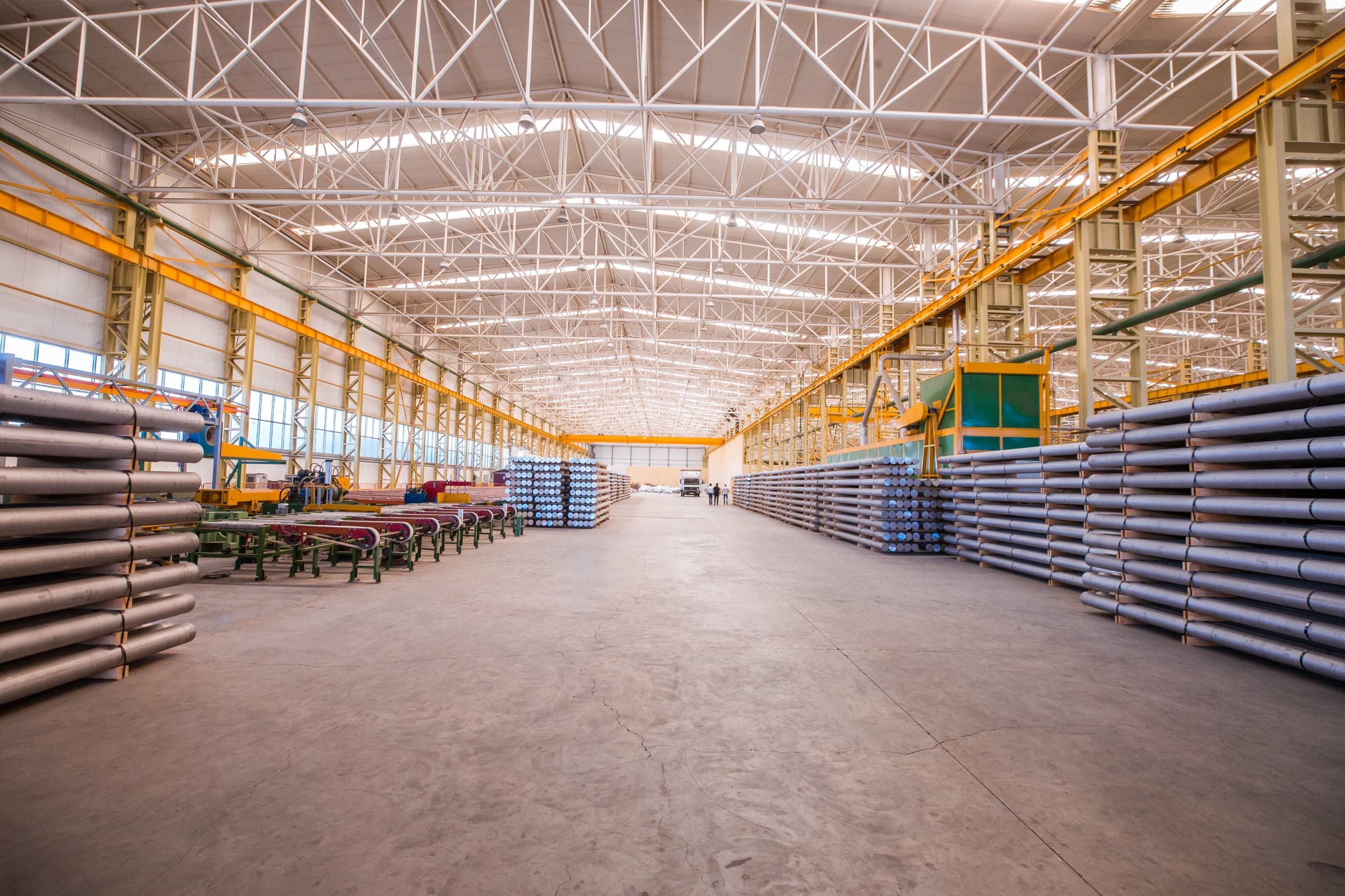 Interior of a large steel-framed industrial warehouse with a long concrete floor and stacked materials